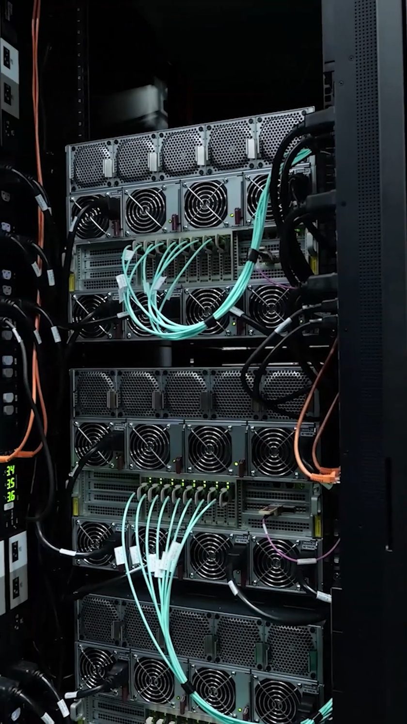 Achterzijde van een GPU-rack tijdens de bouw: twee HGX-klasse nodes bekabeld met optische interconnect, ventilatoren en voedingen zichtbaar.